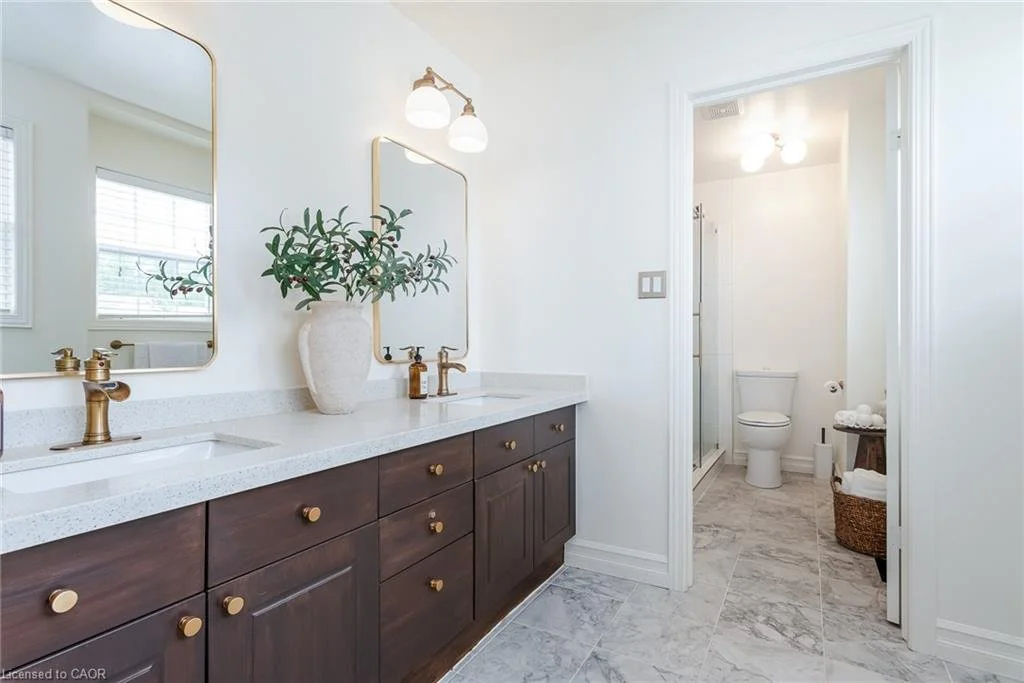 Bathroom with dual sinks, gold fixtures, a large mirror, a vase with greenery, and an adjoining toilet area.
