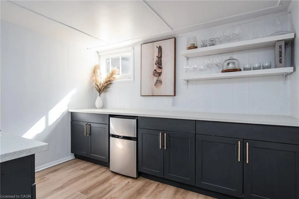 Minimalist kitchen with white paneled walls, black cabinets with gold handles, a small stainless steel refrigerator, open shelves with glassware, and a window with sunlight streaming in.