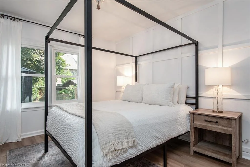 A bedroom with a black four-poster bed, white bedding, a wooden nightstand, and a table lamp. There is a window with white curtains showing trees outside.