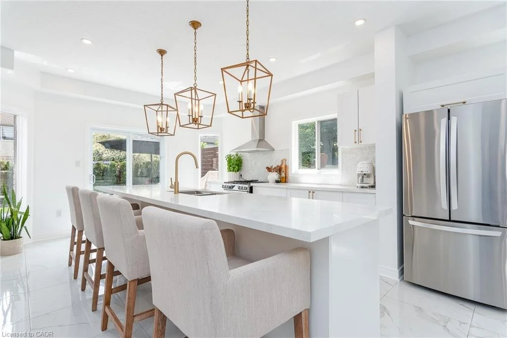 Bright, modern kitchen with white cabinetry, a large kitchen island with beige chairs, stainless steel refrigerator, and natural light from windows and sliding glass door.