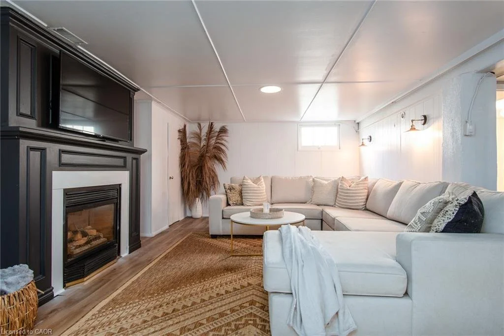 Living room with a white sectional sofa, a round coffee table, a fireplace with a black mantel, a small window, wall-mounted lights, a large plant, and a woven rug.
