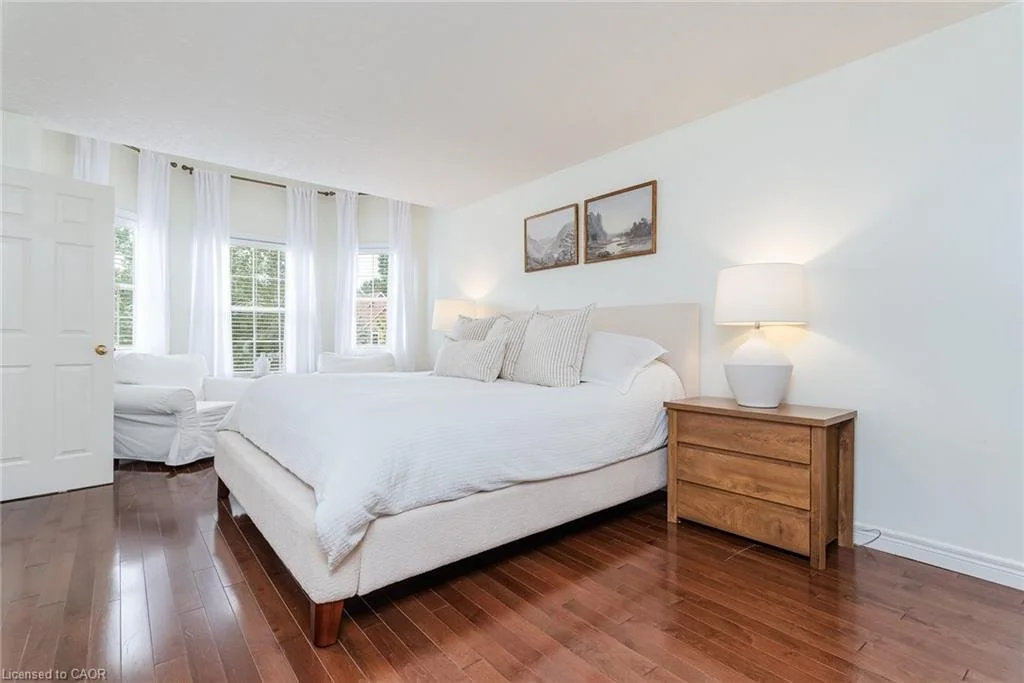 A bright bedroom with white walls, a large bed with white bedding and pillows, wooden side table with a lamp, a white armchair, and multiple windows with white curtains.