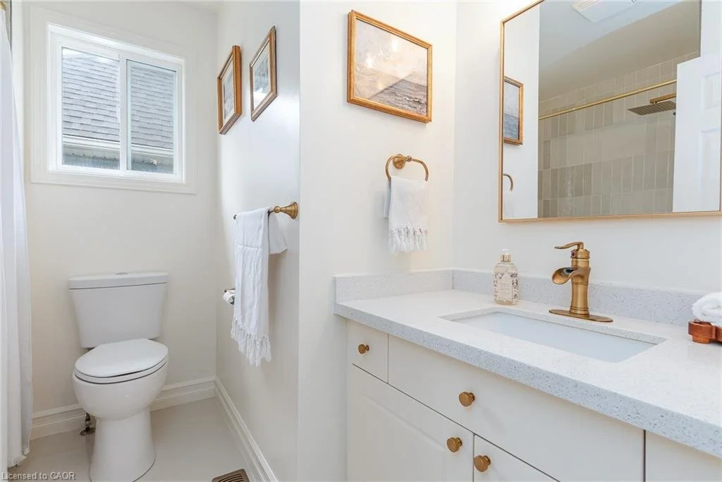 A clean white bathroom with a toilet, a white vanity with a gold faucet, a large mirror, a window, framed artwork on the walls, and a towel hanging on a gold towel ring.