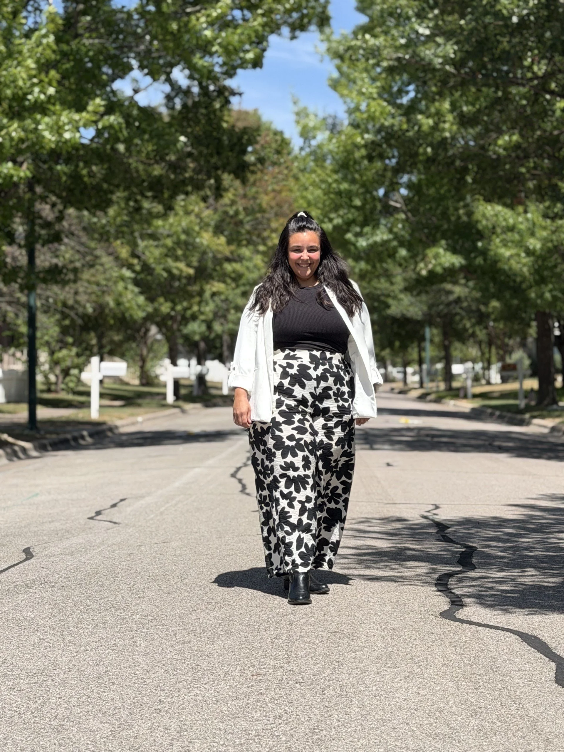 A woman walking on a paved street lined with trees, smiling, wearing black and white patterned pants, a black top, a white blazer, and black boots, with a blue sky overhead.