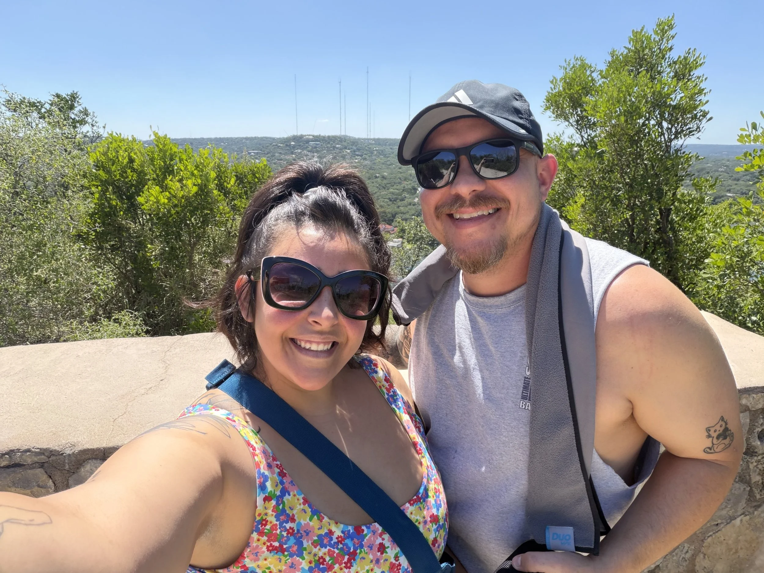 Smiling woman and man taking a selfie outdoors on a sunny day, with a background of green trees, hills, and radio towers in the distance.