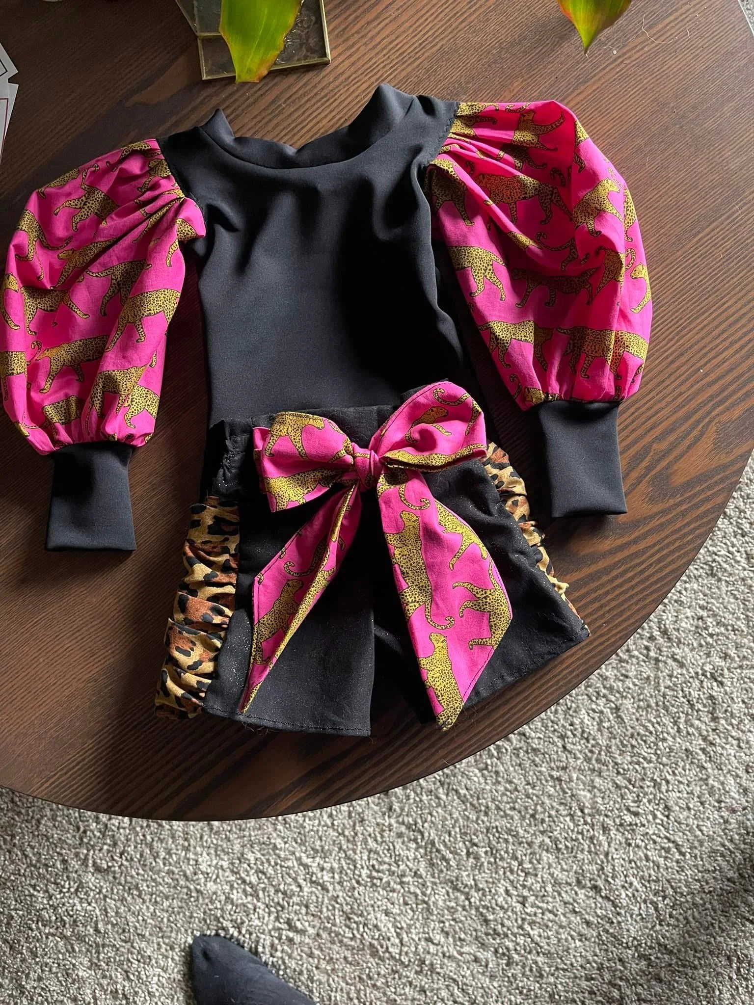 A black hoodie with bright pink and yellow leopard print sleeves and a matching leopard print bow on the front, placed on a wooden table.