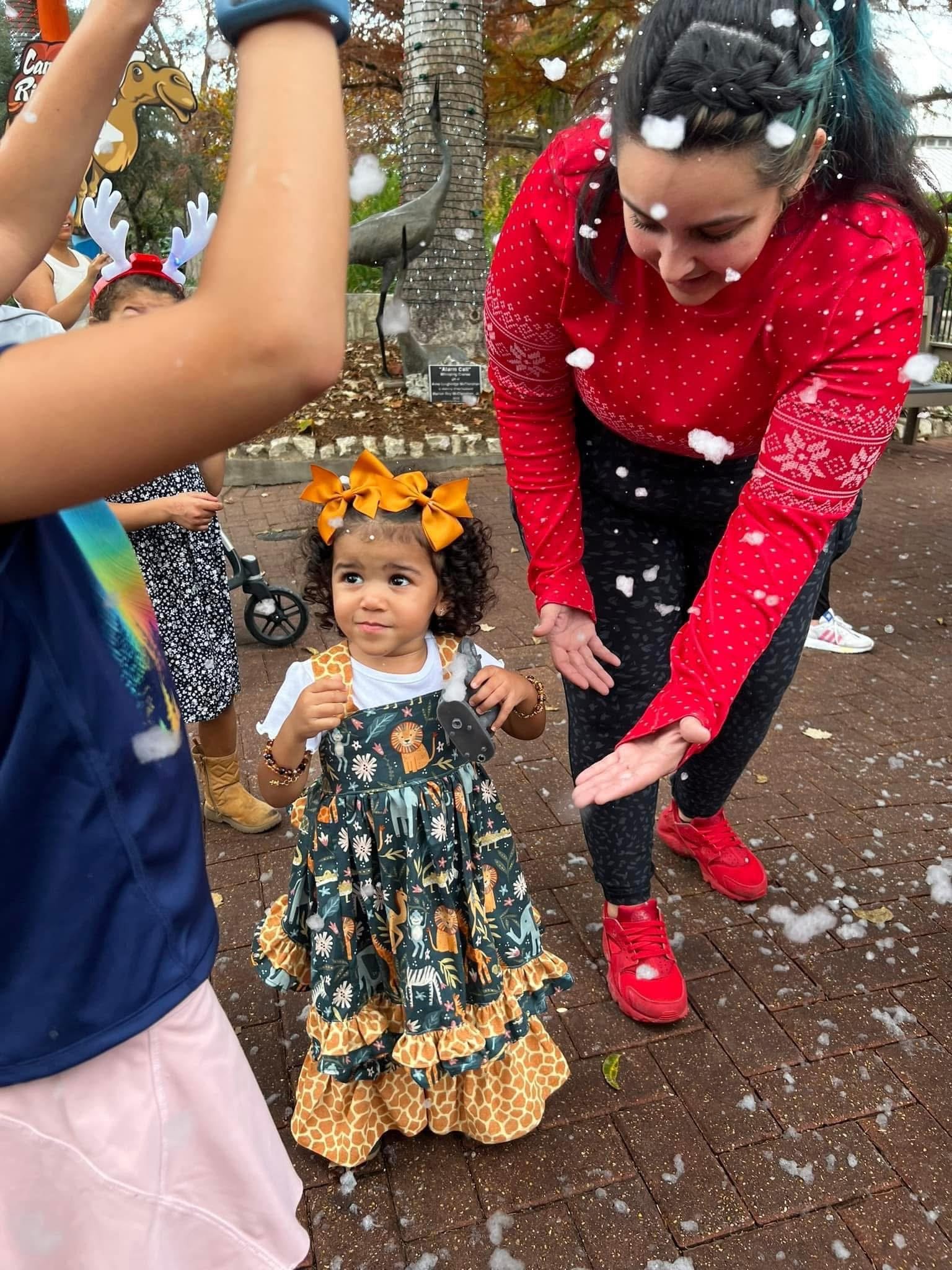 A young girl in a giraffe-themed dress and large orange bow on her head standing outdoors surrounded by other children, one in antlers headband, and an adult in a red holiday sweater. There is snow or foam on the ground and in the air, with trees and a sculpture in the background.