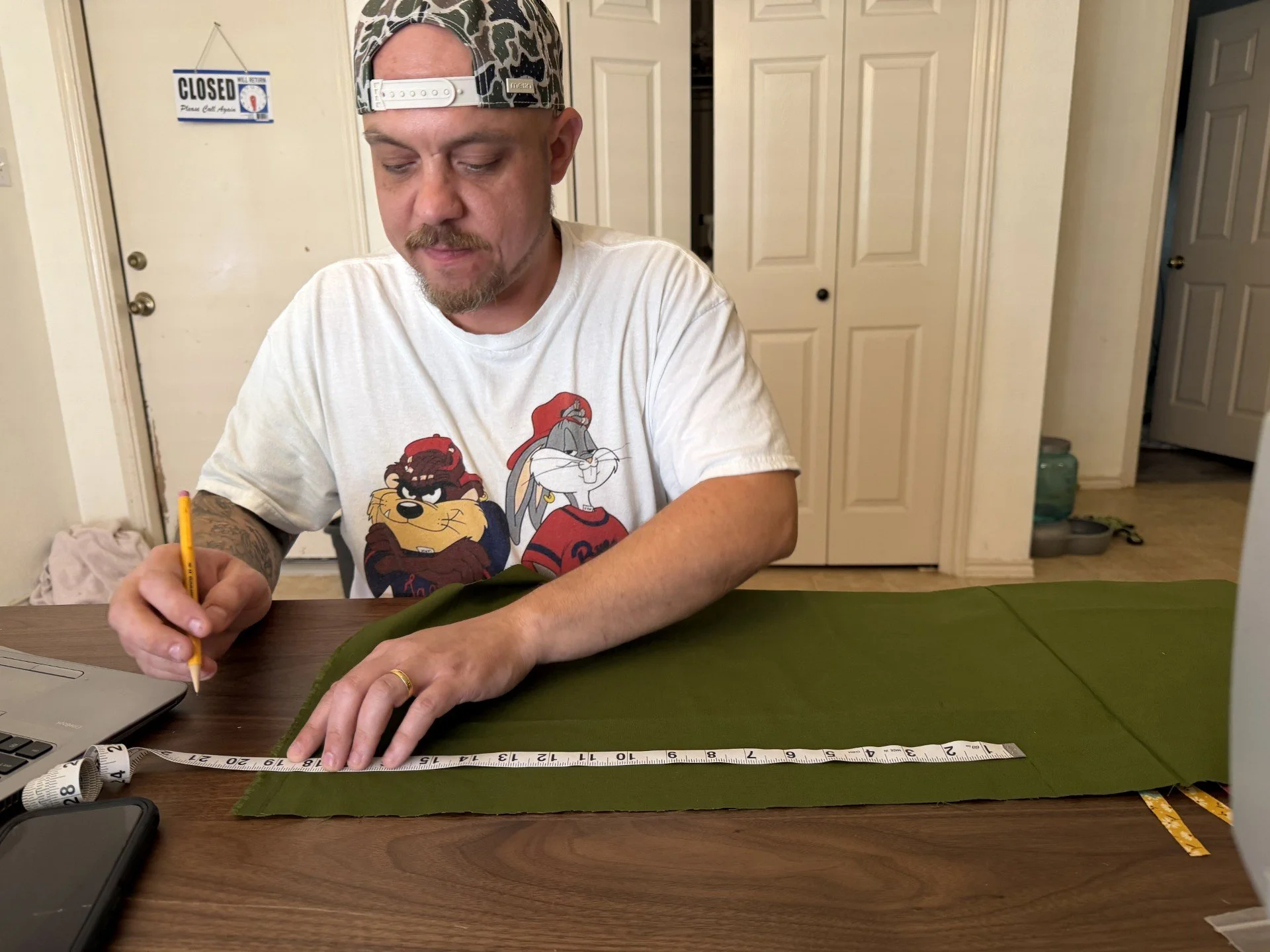 Man sitting at a table, measuring fabric with a yellow tape measure. He is wearing a white t-shirt with cartoon characters and a camouflage baseball cap worn backwards.