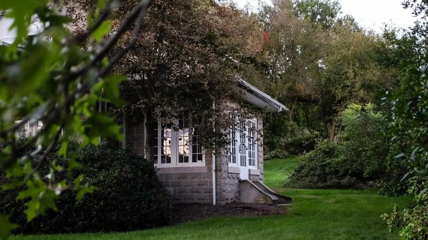 A house with brick and white siding, surrounded by green trees and bushes, with a grassy yard in the foreground.