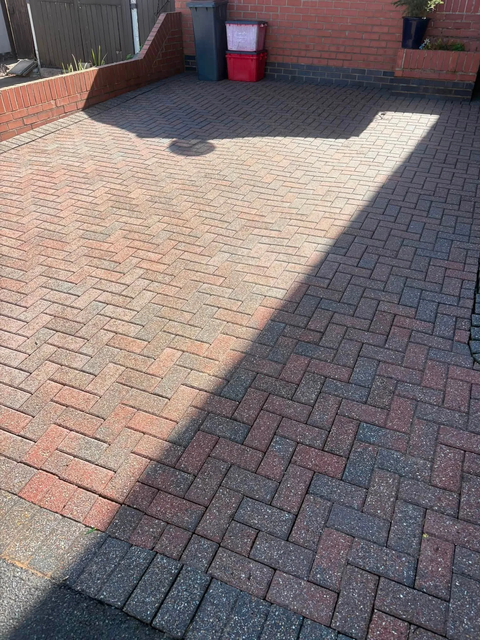 A brick-paved driveway next to a brick house wall, with a shadow cast over part of the pavement. Two stacked red and white containers and a dark green trash bin are placed against the brick wall. A black planter with some plants is mounted on the wal