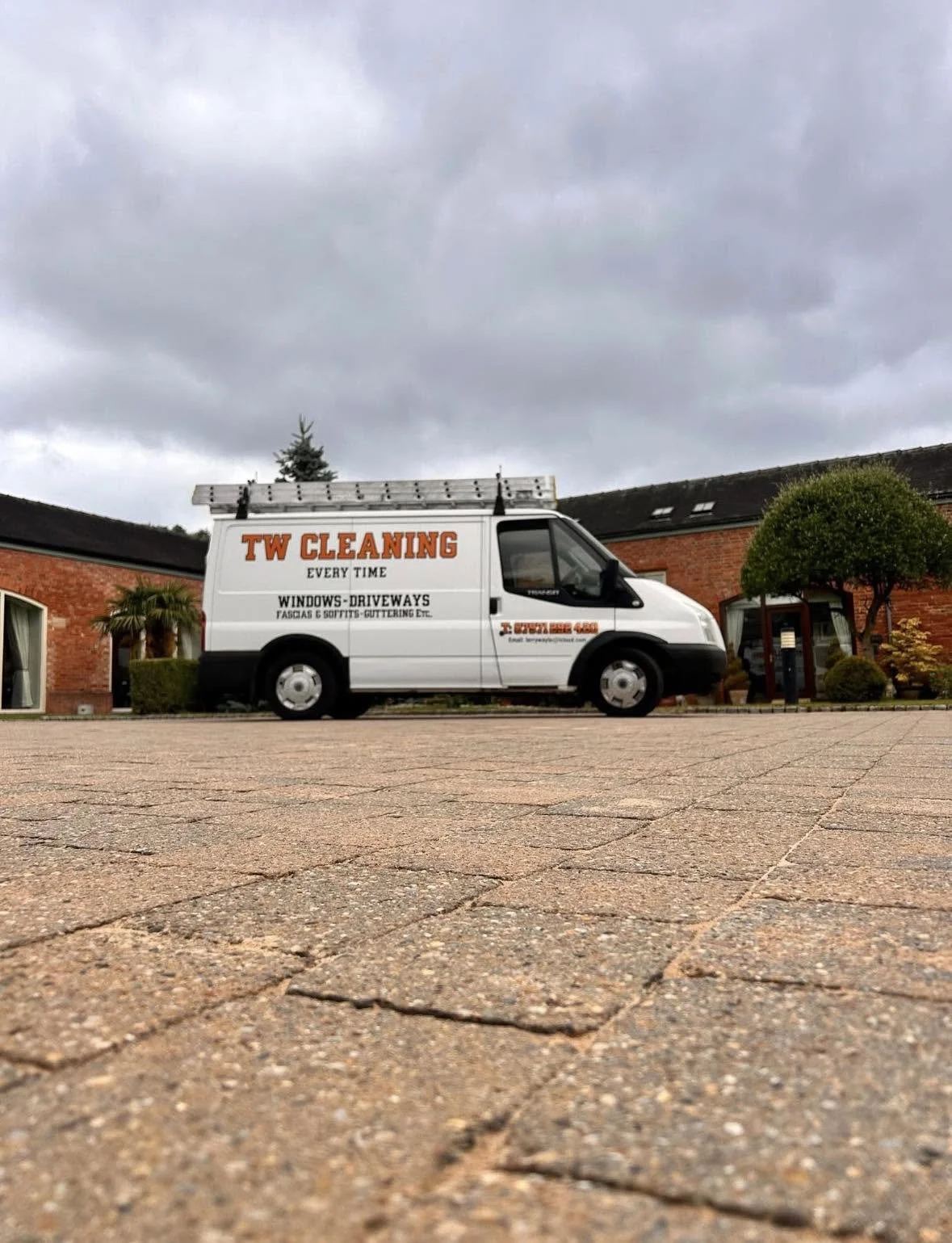 White company van with orange and black text parked on a brick driveway in front of a brick house with greenery and trees.