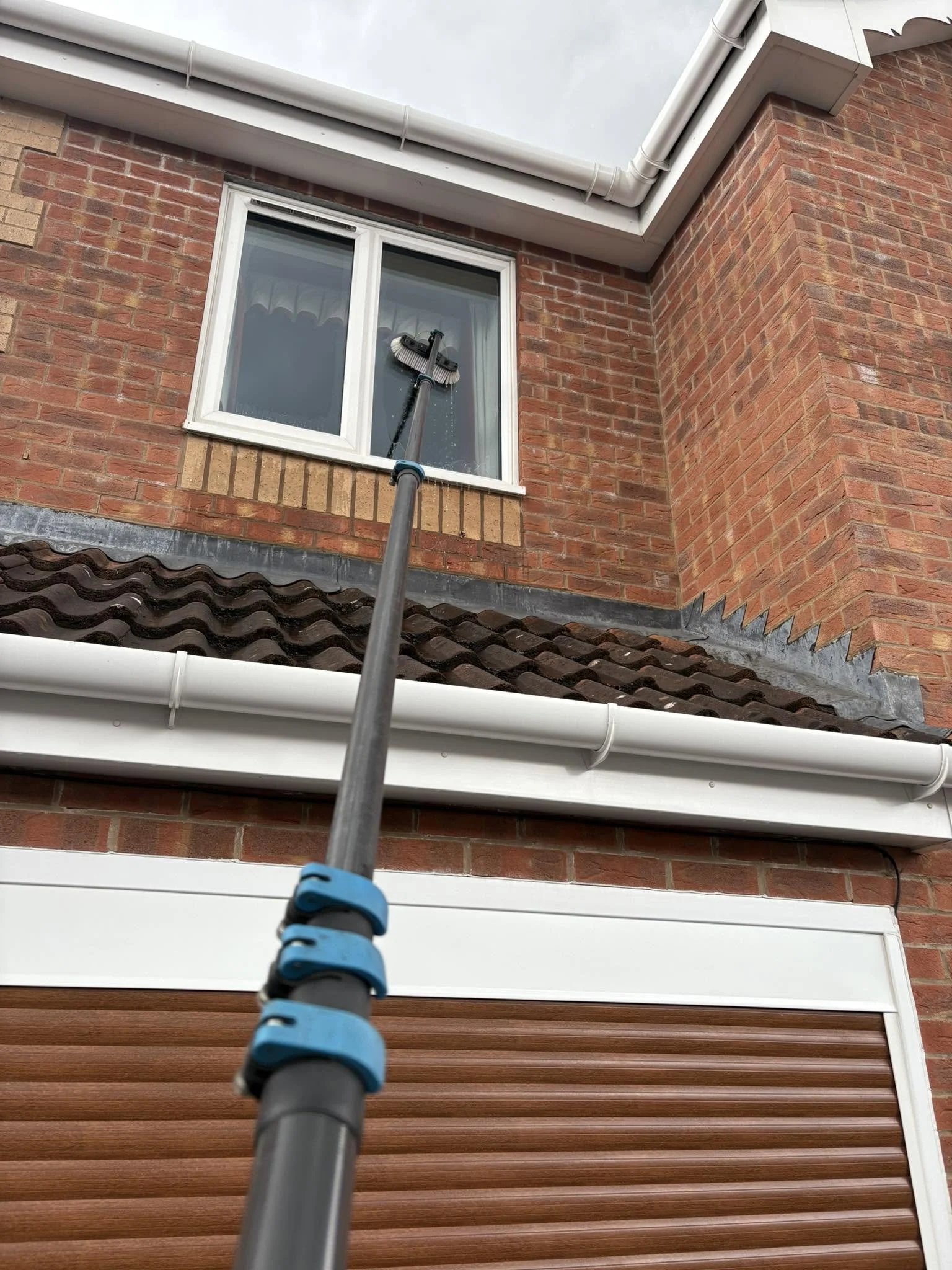 A person cleaning a second-story window on a brick house with a long-handled squeegee, during daytime with overcast sky.