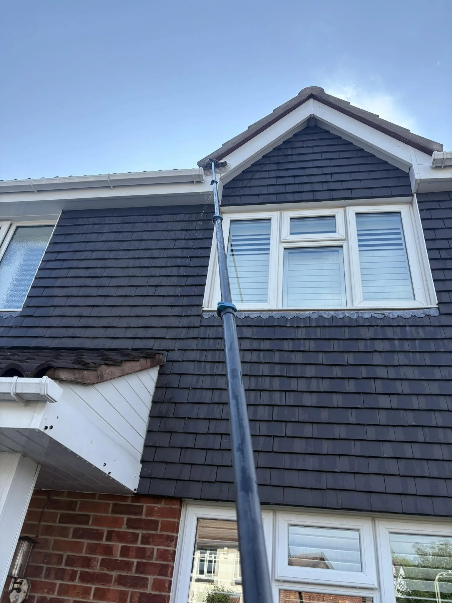 Close-up of the upper part of a house with dark shingles, a window with blinds, and a gutter, with a ladder or pole visible in the foreground, under a blue sky.