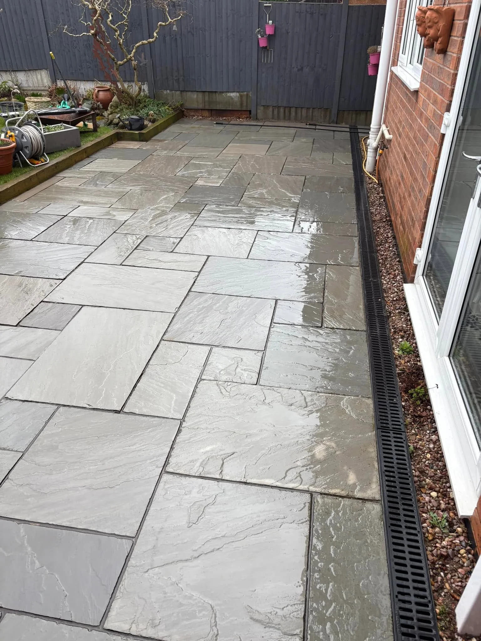 Clean outdoor patio with gray stone tiles, brick wall on the right, black wooden fence in the background, and gardening tools and plants on the left. Water on the tiles indicates recent cleaning.