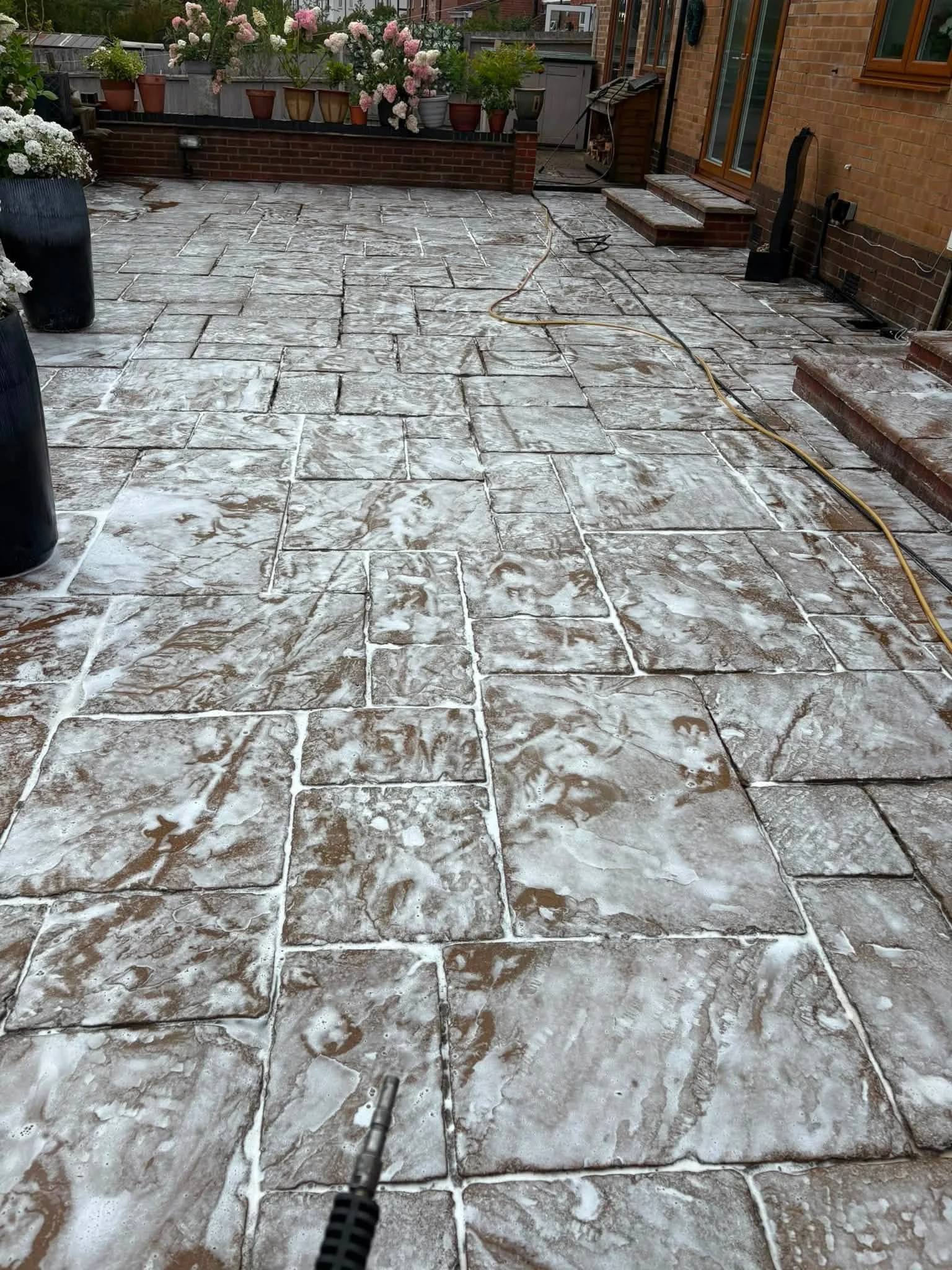 A backyard patio with large stone pavers that are being pressure washed, visible soap and water on the surface. The background features potted plants with pink and white flowers, a brick house with a sliding glass door, and some gardening tools and h