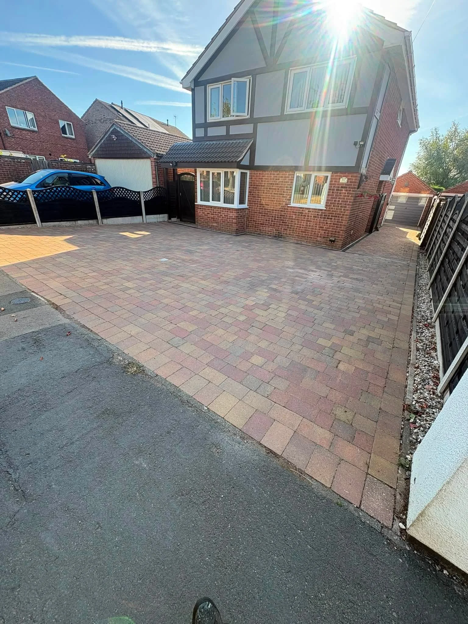 A paved driveway in front of a house, with neighboring houses visible, a car parked on the left, and the sun shining brightly, creating a lens flare.