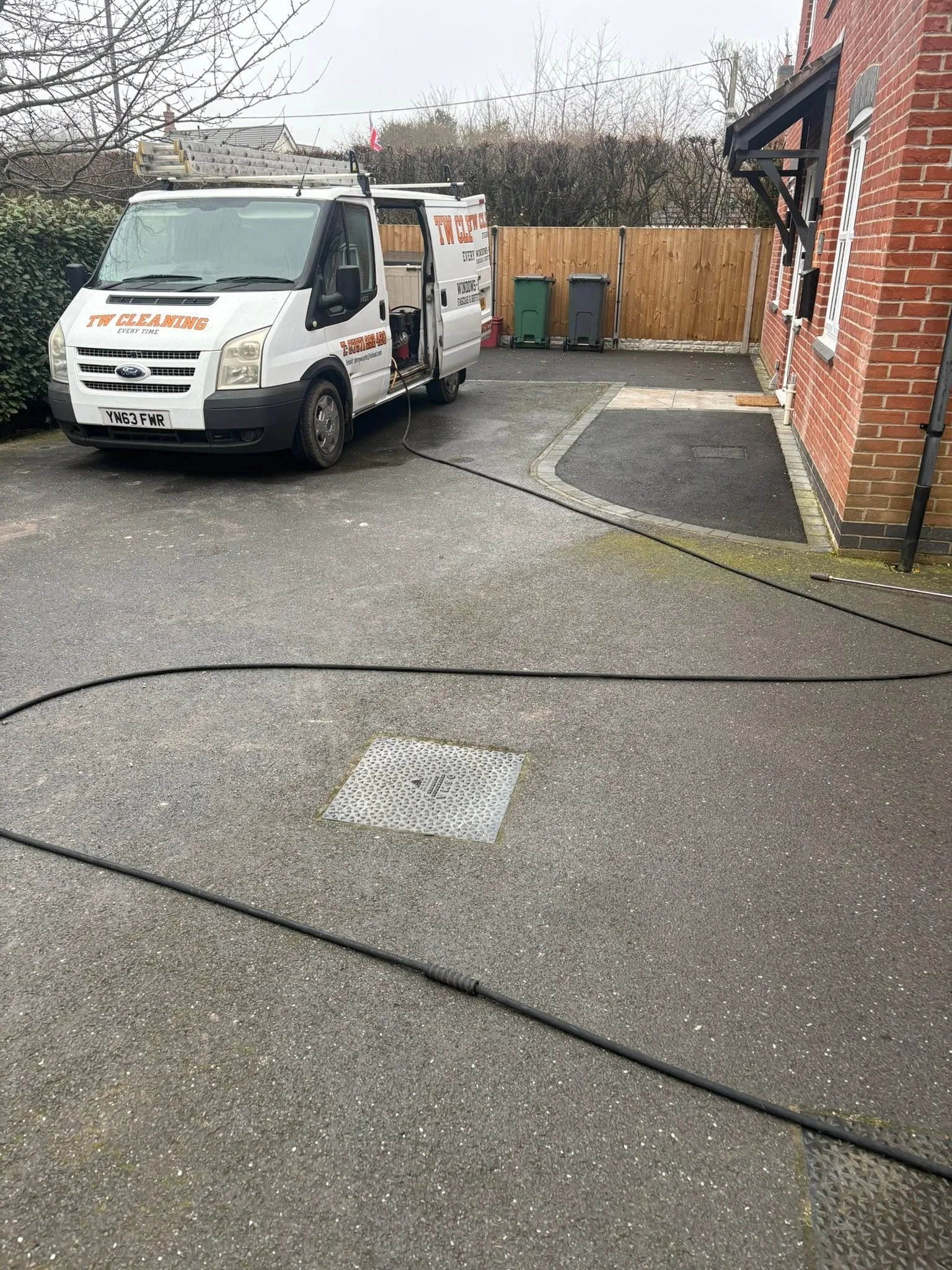 A cleaning service van parked in a driveway, with cleaning equipment connected to it, near a brick house, with trash bins and a wooden fence in the background.