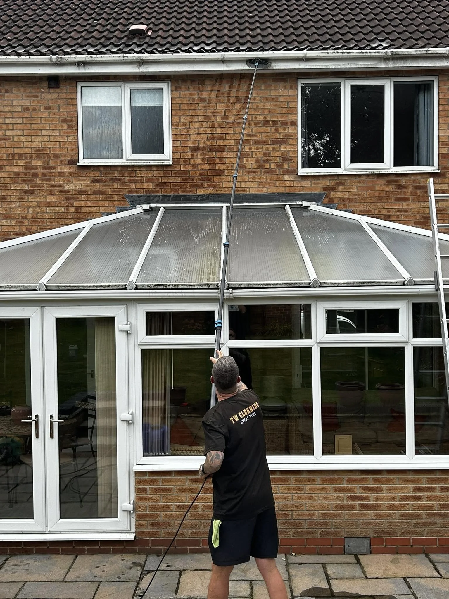 A man cleaning the exterior glass of a sunroom or patio enclosure attached to a brick house. He is standing on a paved surface and wearing a black T-shirt and shorts, holding a cleaning tool connected to a long hose.