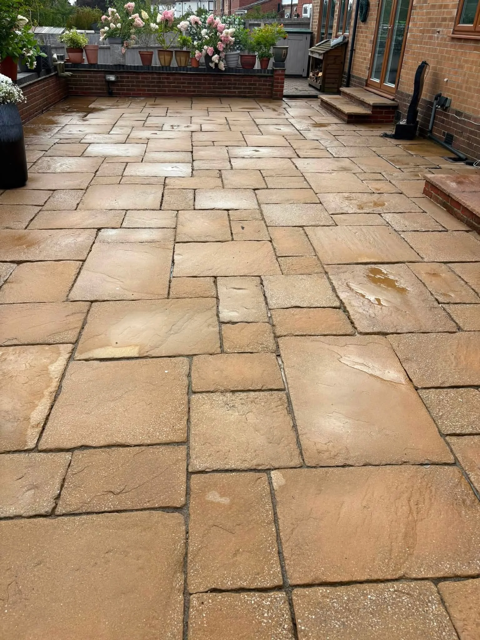 A wet stone patio with rectangular and irregularly shaped tan and beige stones, bordered by a brick wall and potted plants with blooming flowers.