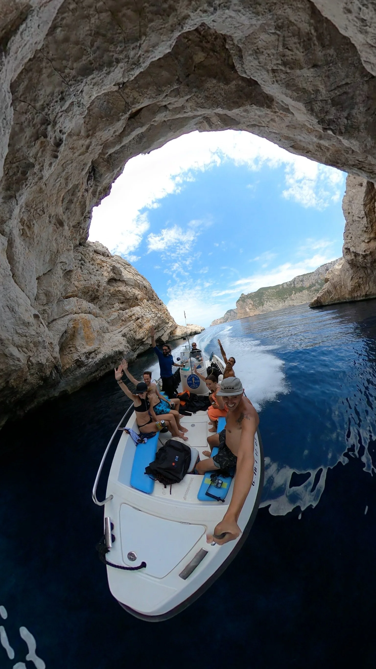 Group of people on a boat passing through a rocky waterway with a natural rock arch overhead, blue sky, and distant cliffs visible.