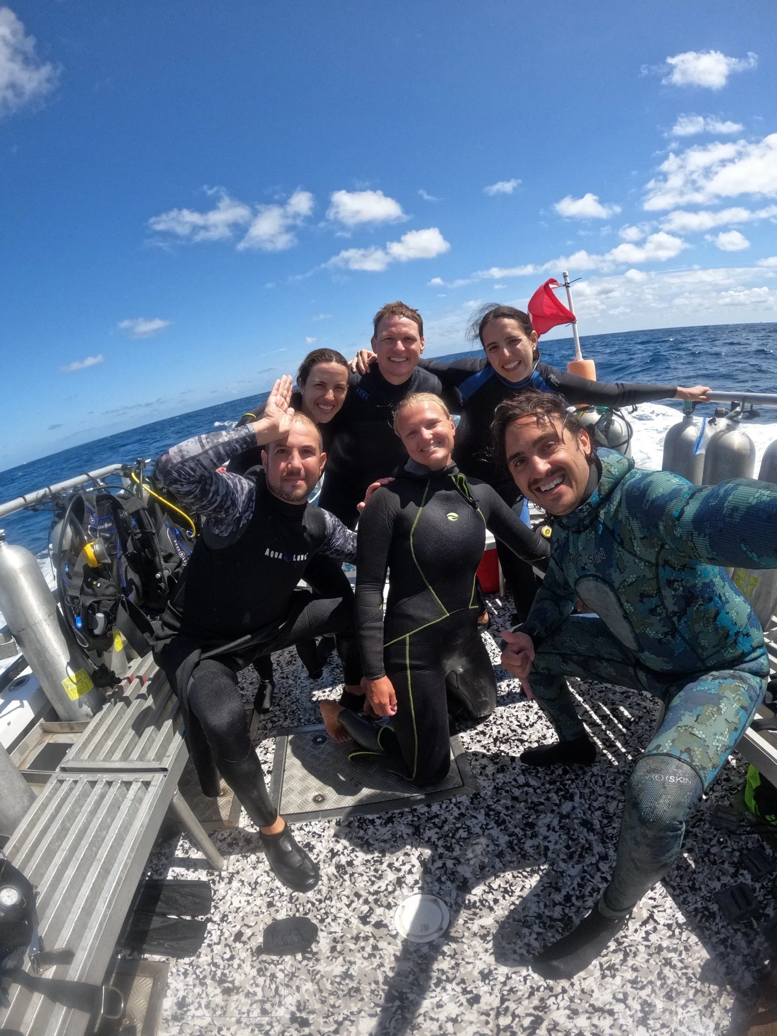 A group of six people in wetsuits on a boat at sea, smiling and taking a selfie, with a blue sky, some clouds, and the ocean in the background.