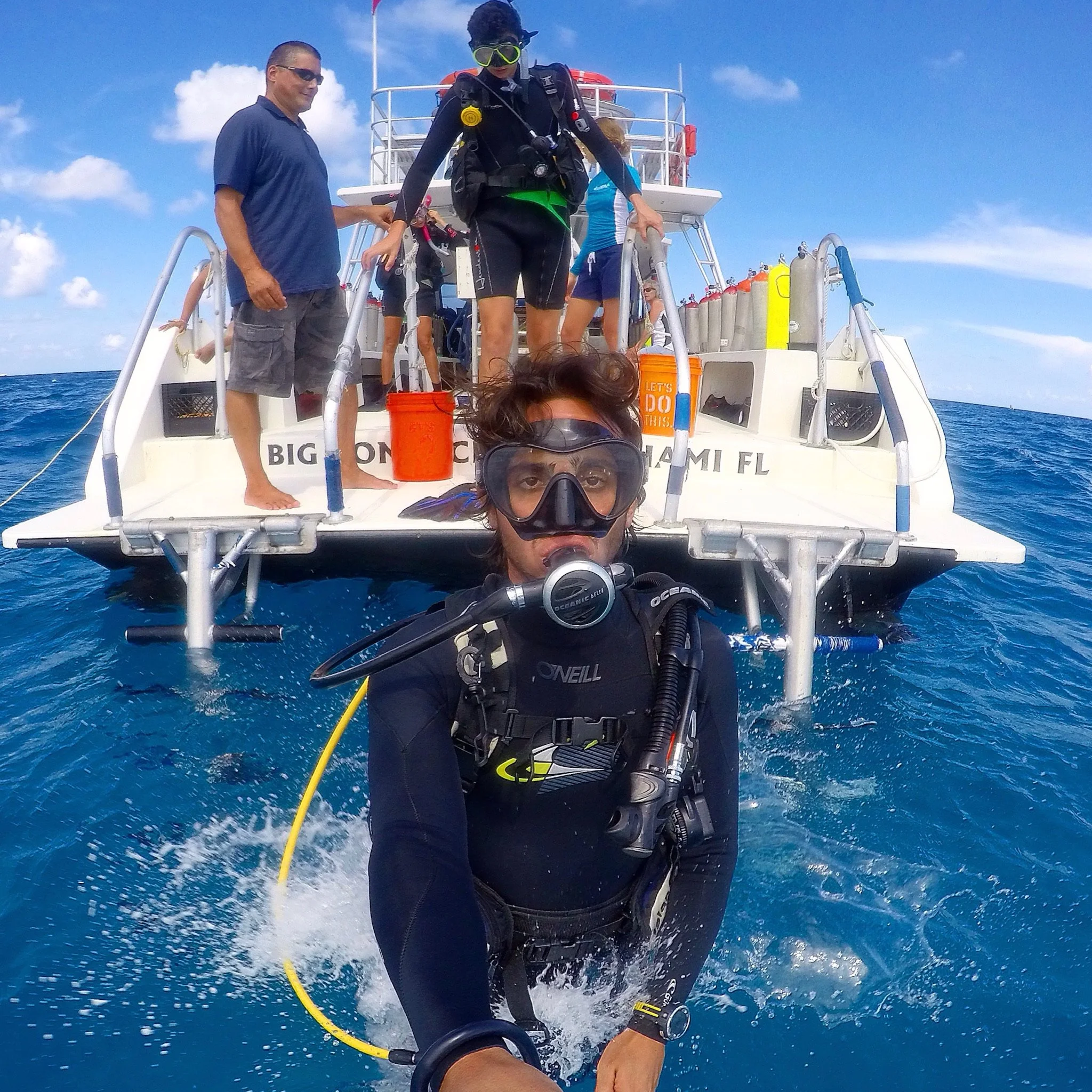 Person wearing scuba diving gear riding on the back of a boat in the ocean, with a man standing beside them and other people in the background on the boat, under a partly cloudy sky.