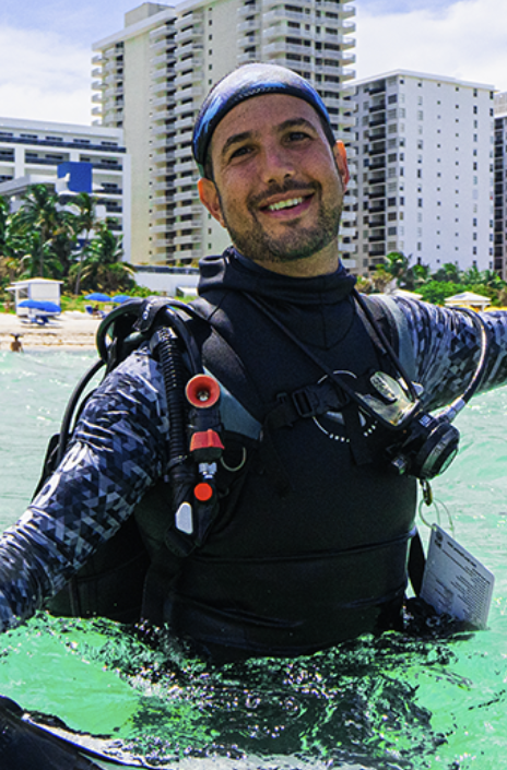 A smiling man in scuba gear standing in shallow ocean water with high-rise buildings and beach umbrellas in the background.