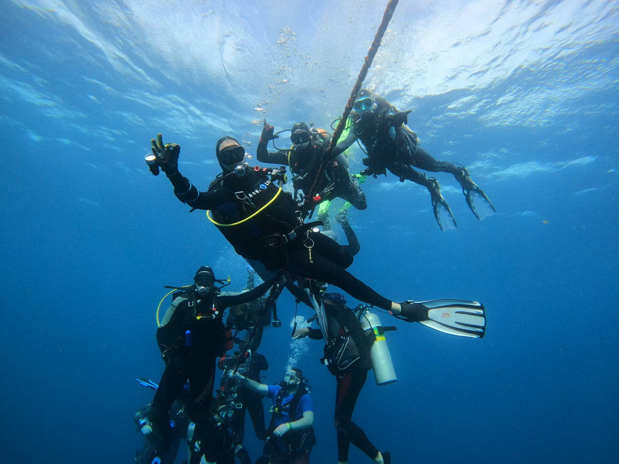 Group of scuba divers underwater holding onto a buoy line, wearing diving gear including masks, wetsuits, and fins, in clear blue water.