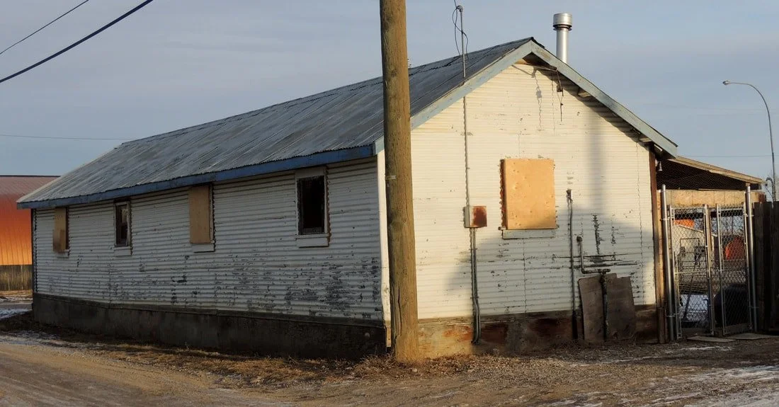 An old, weathered white house with boarded-up windows and a corrugated metal roof, situated on a dirt road, with a utility pole in front.