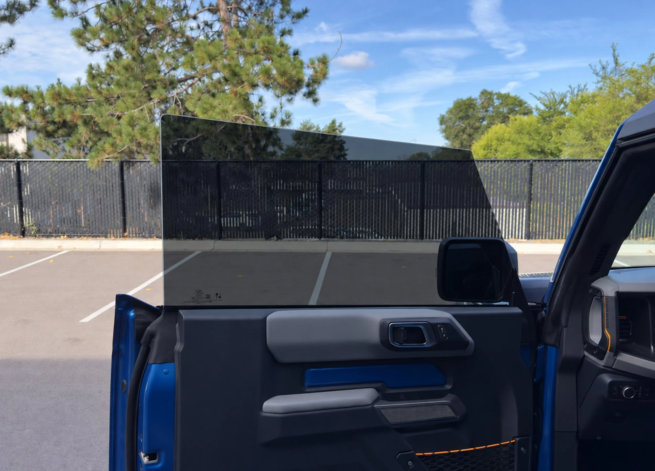 Vehicle window with a parking area, fence, and trees visible behind it.