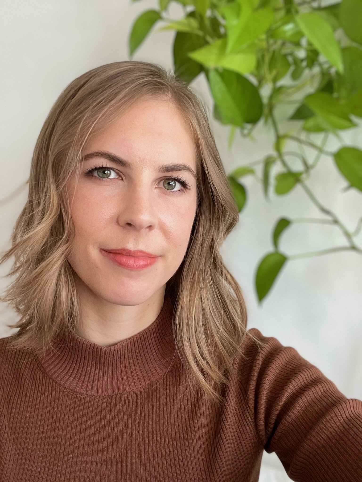 A young woman with wavy light brown hair and green eyes taking a selfie in front of a white wall and green leafy plant, wearing a brown ribbed sweater.