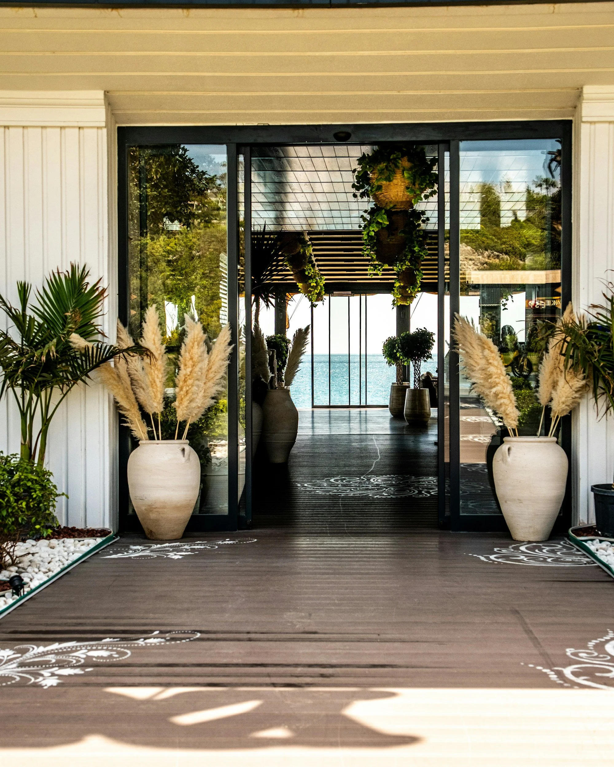 View through glass doors to an outdoor area with water, plants, and a wooden pergola. Potted plants decorate the entrance, and decorative white patterns are on the wooden floor.