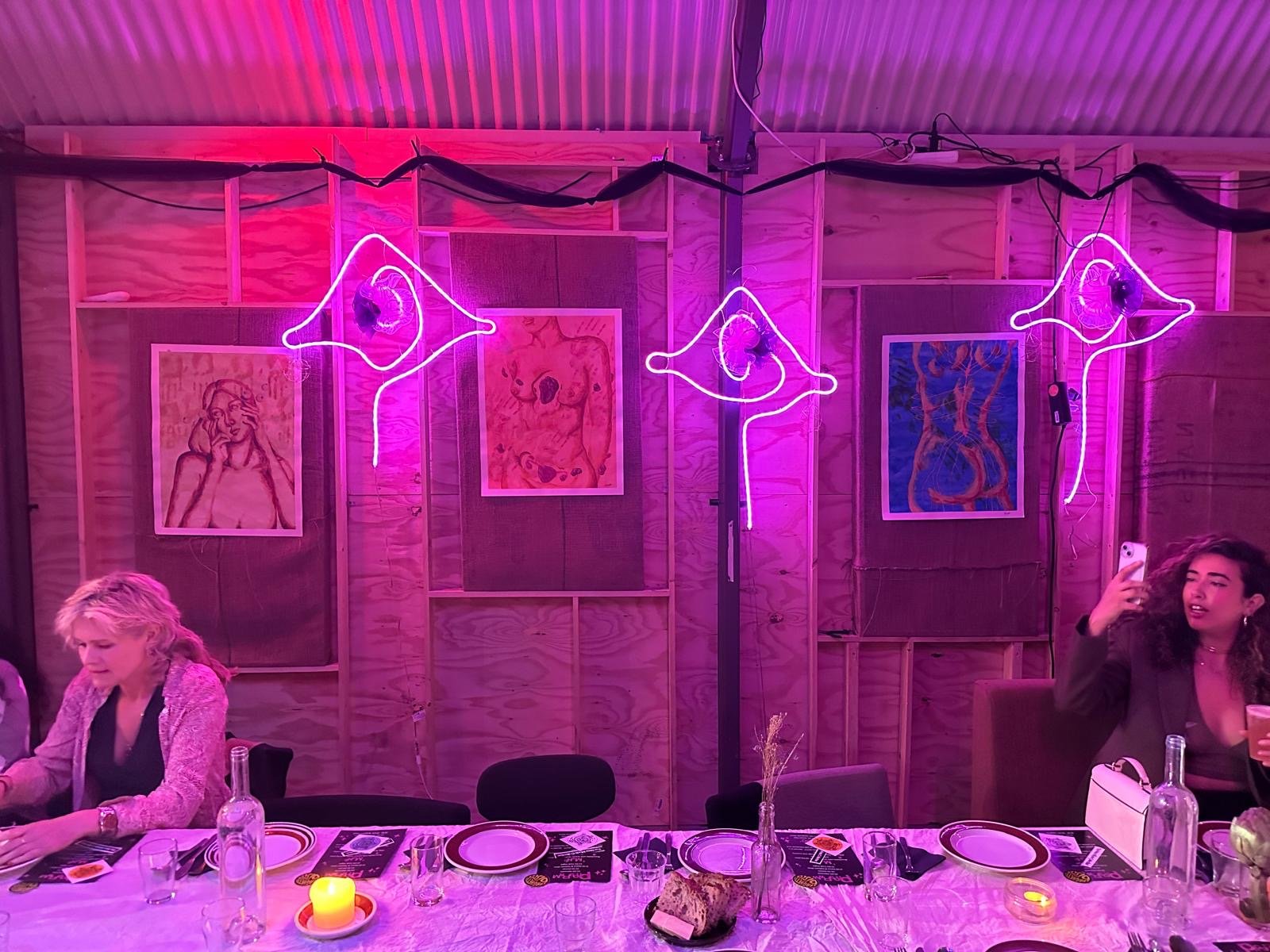 A restaurant or event space with a wooden wall decorated with framed art and neon lights shaped like lips and a face. People are sitting at a table, one woman is taking a selfie, and the table is set with plates, glasses, and a candle.