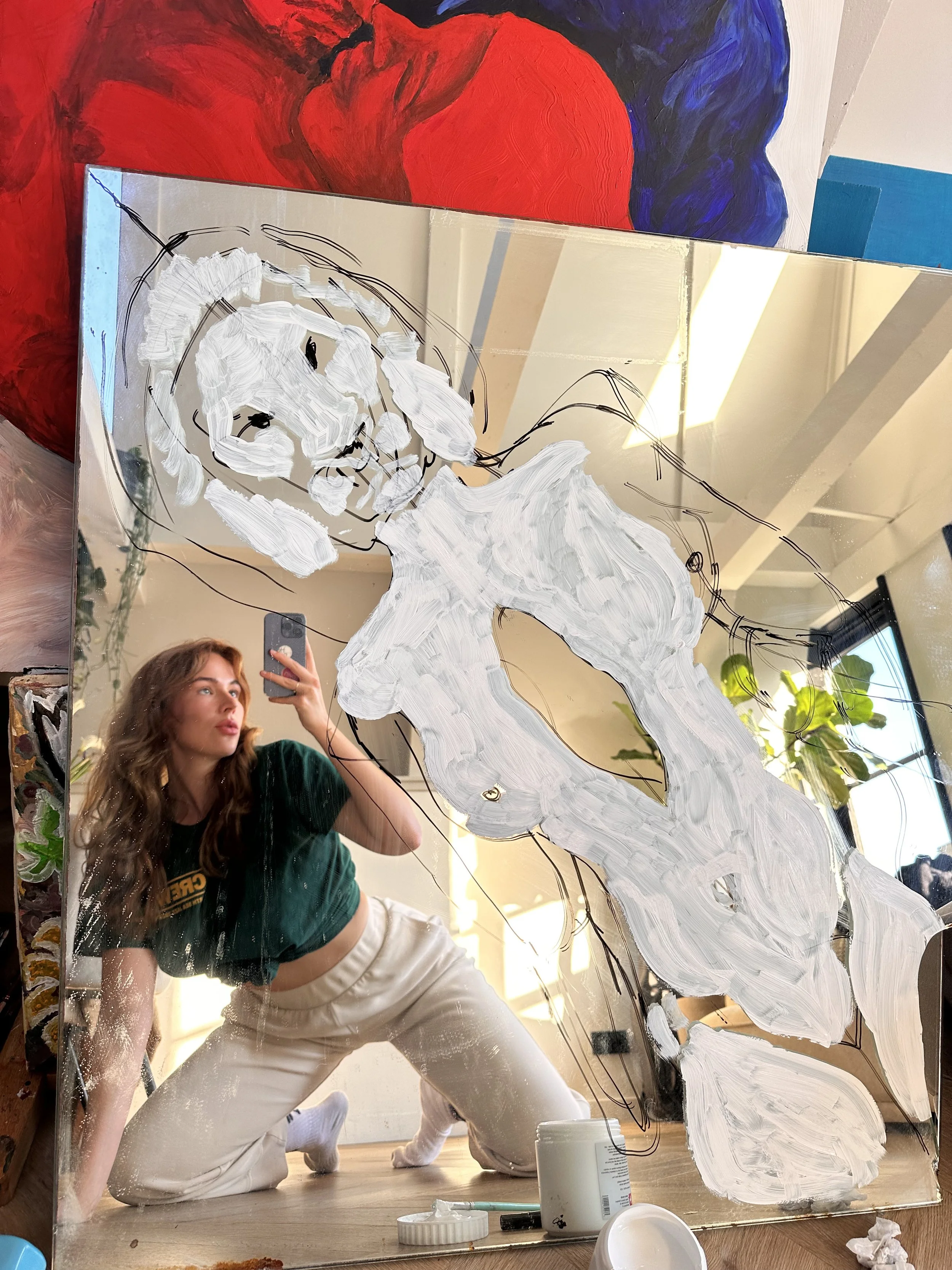 A young woman with long wavy hair taking a mirror selfie with her phone. She is bending down, wearing a green cropped sweatshirt and light-colored pants. The mirror has white paint and black lines drawn on it, creating an abstract design. There are art supplies like a white container, a pen, and a lid placed on the wooden floor in front of the mirror. In the background, there are colorful abstract paintings and a window with sunlight streaming in.