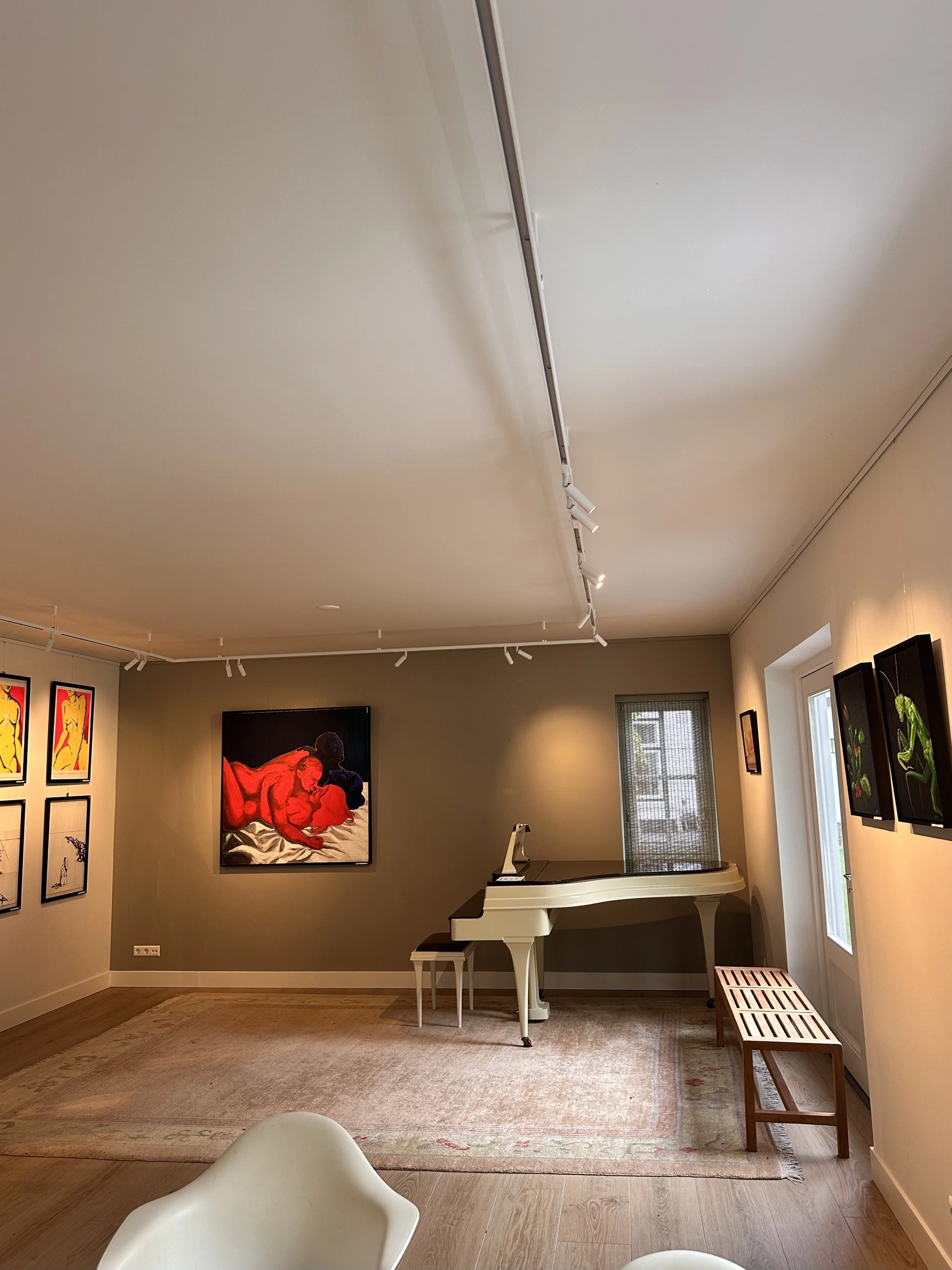 Interior of an art gallery with paintings on the wall, a white piano, and seating area, illuminated by ceiling track lights.