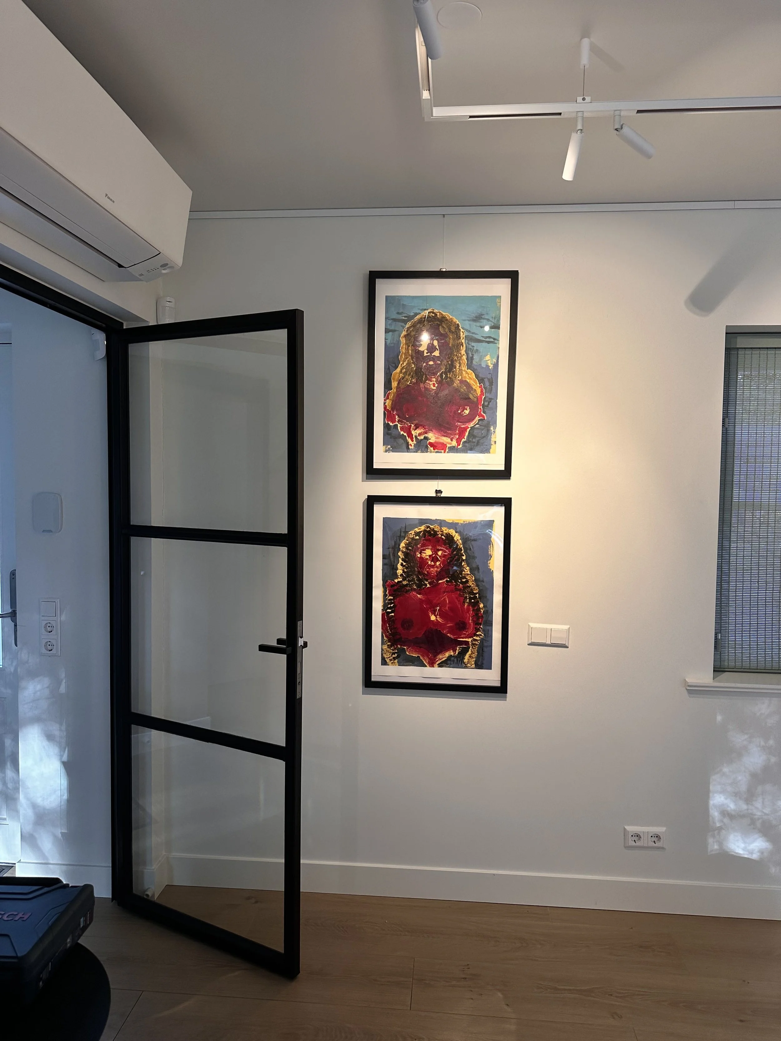 Interior of a room with white walls and wooden flooring, featuring two framed colorful portraits of women with long hair, hanging vertically on the wall. There is a black-framed glass door on the left and a window with blinds on the right.