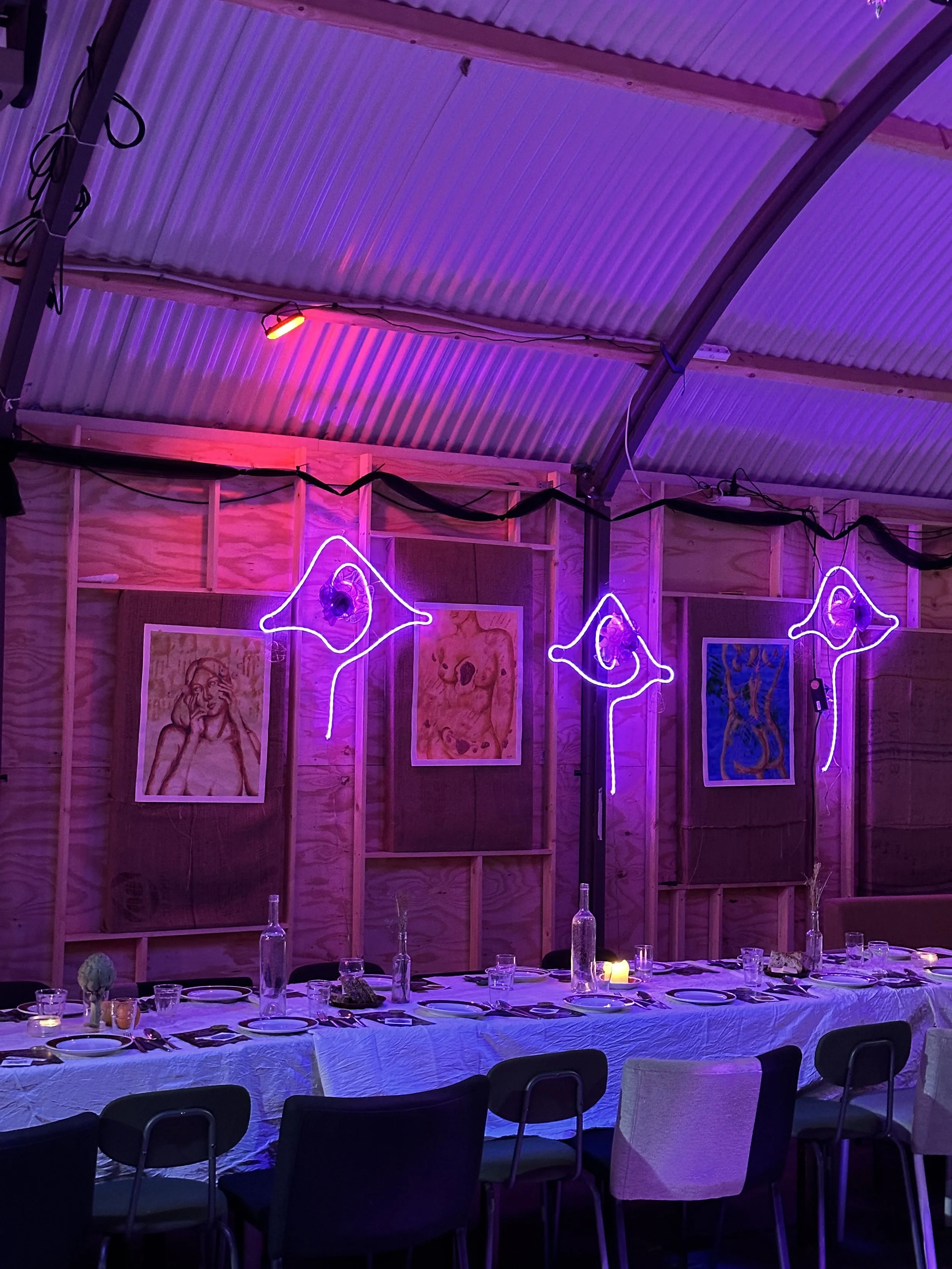 A decorated indoor dining area with a table set for a meal, surrounded by chairs. The area has purple lighting, with neon star-shaped lights and framed artwork on the wooden walls. Bottles, glasses, and tableware are arranged on the table.
