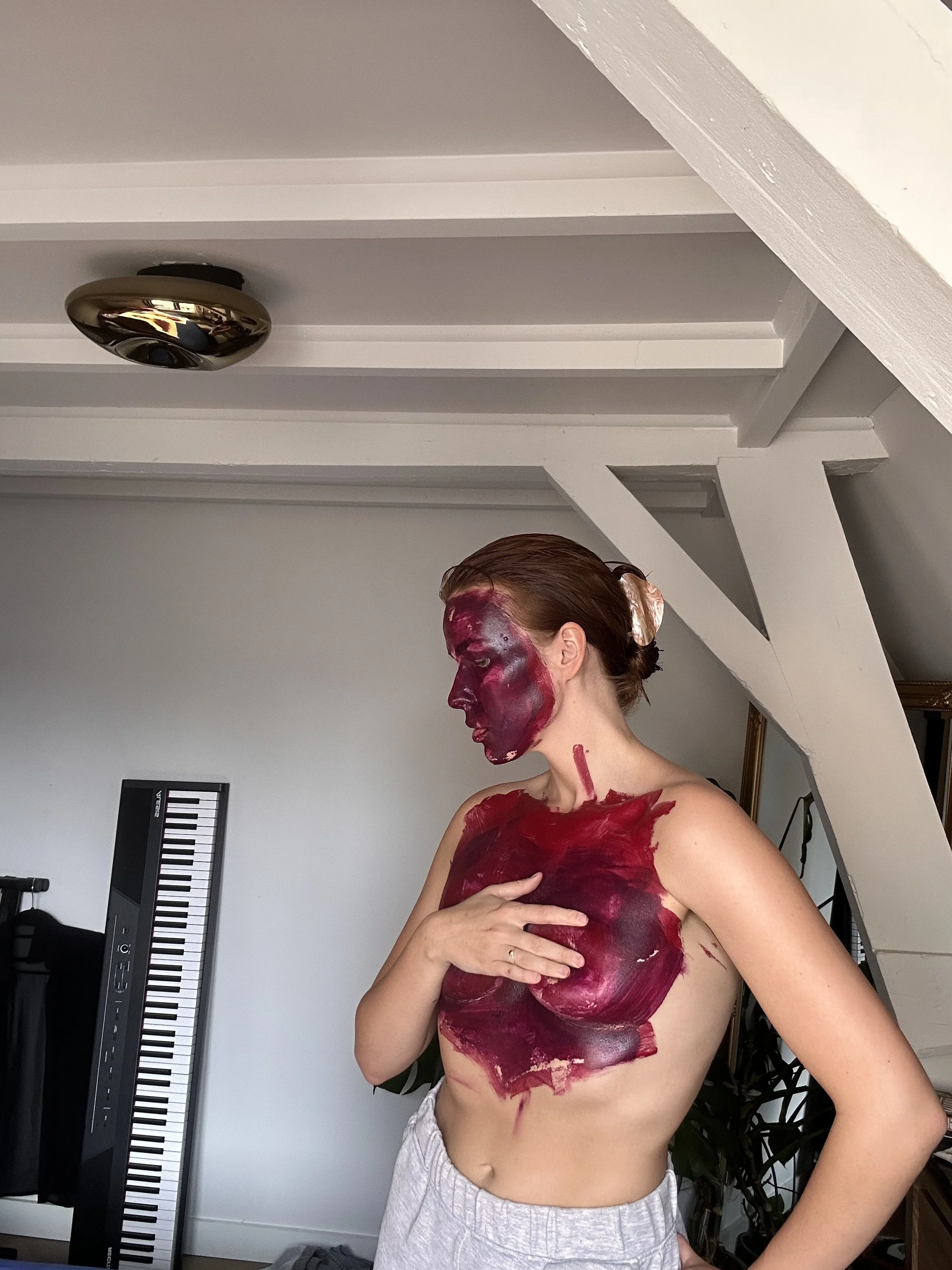 A woman with red face and body paint, wearing a white hair scrunchie and light-colored shorts, standing in a room with a keyboard and framed pictures, pressing her right hand against her chest.
