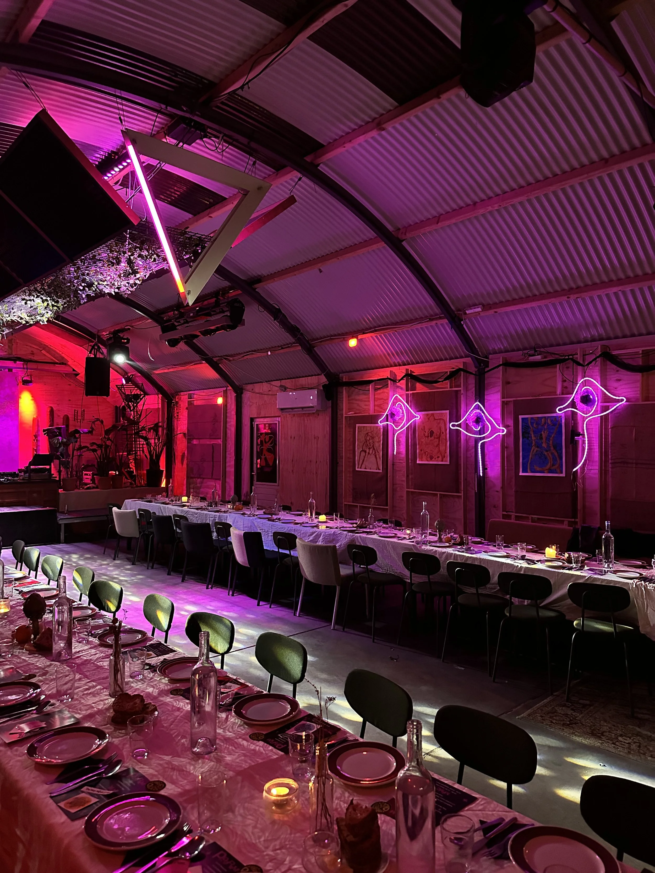 An indoor event space with a long dining table, set with plates, glasses, utensils, and candles, under pink and purple lighting, with art on the walls and neon eye-shaped signs.