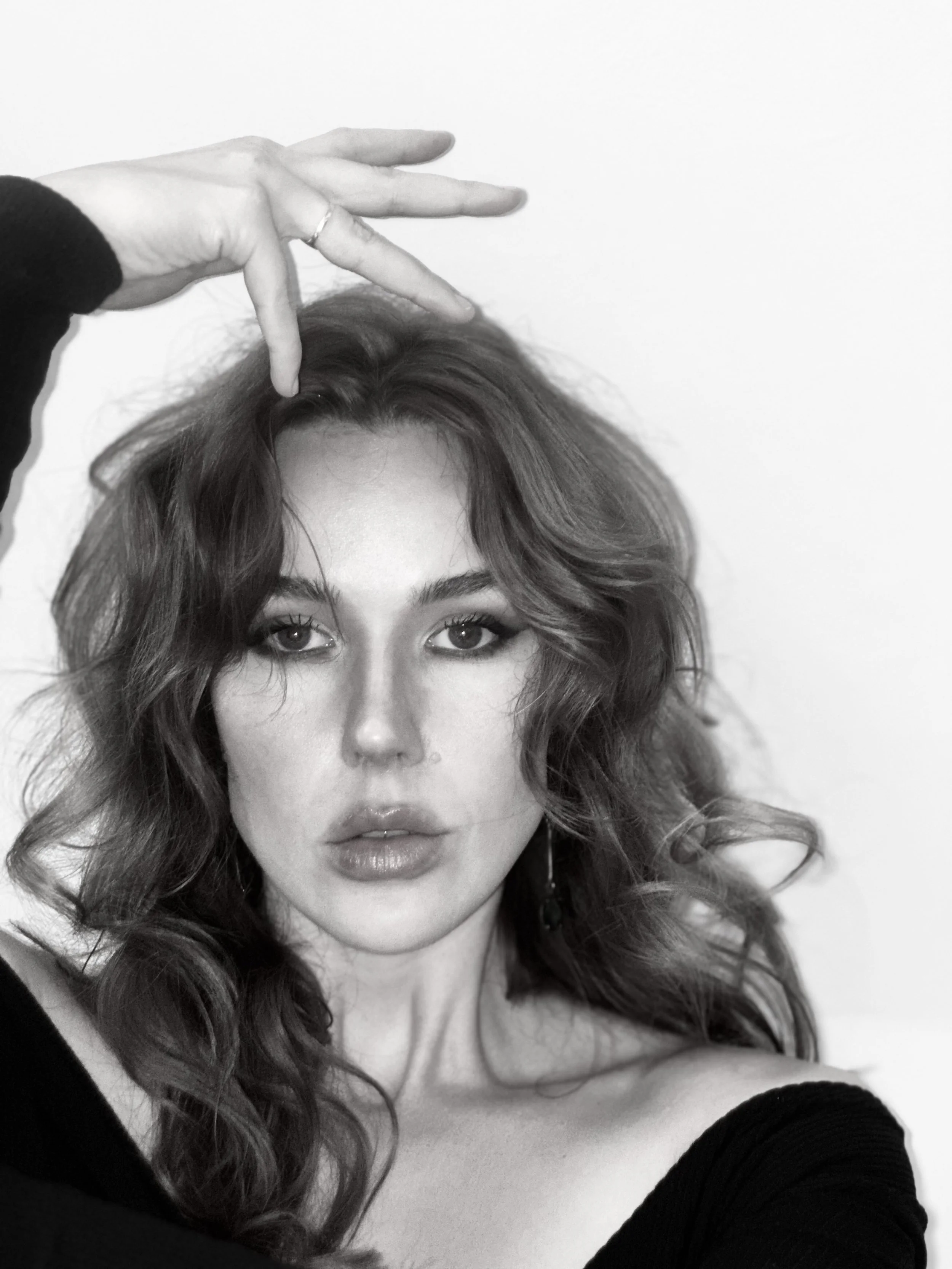 A black and white photo of a woman with wavy hair, eye makeup, and lipstick, looking directly at the camera with her lips slightly parted. She is holding her hand above her head with her fingers positioned as if she is balancing or posing.