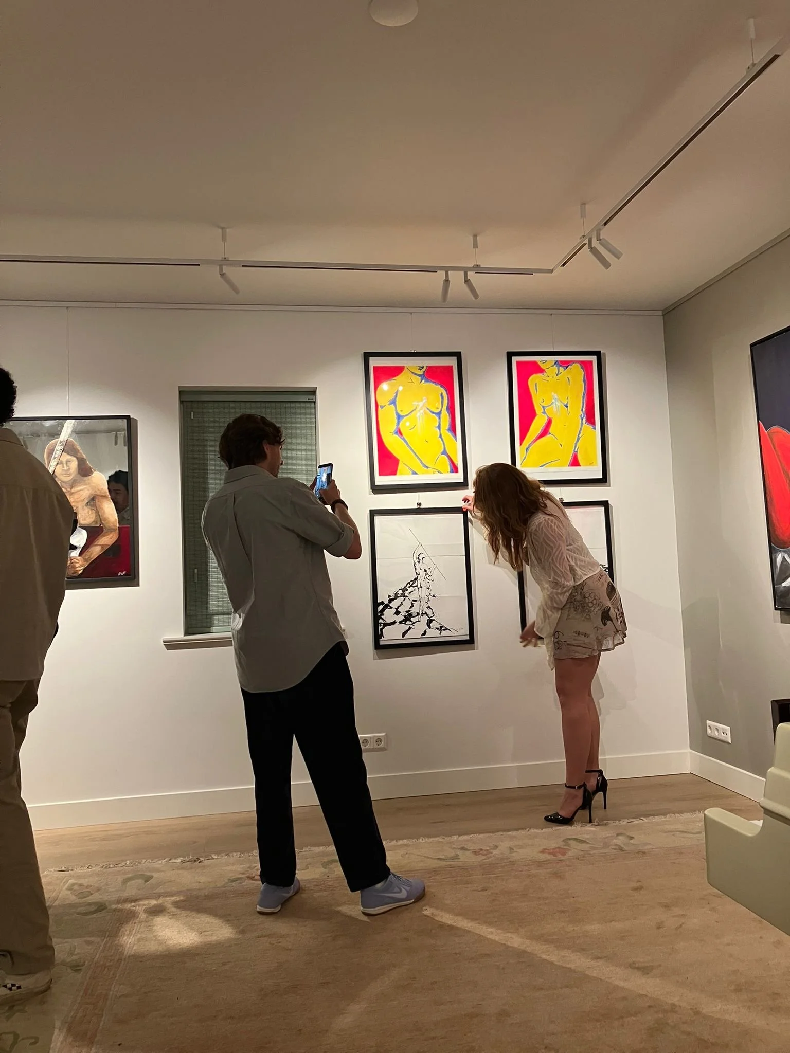 People at an art gallery observing and taking pictures of artwork on the wall, including two colorful portrait paintings, with some visitors engaged in conversation.