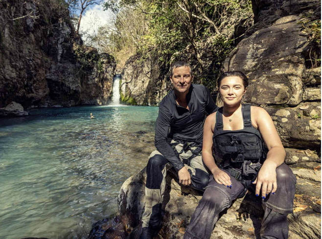 Two people sitting on rocks near a river with a small waterfall in the background, surrounded by trees and rocks.