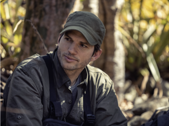 Actor Ashton Kutcher outdoors in a wooded area, wearing a green cap and dark jacket, looking thoughtfully to the side. Promoting the show Running Wild with Bear Brylls