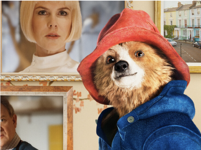 A dog wearing a red hoodie and blue jacket, with portraits of a woman and a man hanging on the wall behind it.