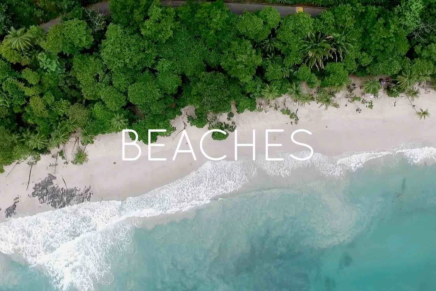 Aerial view of a beach with white sand, turquoise water, and lush green trees along the shoreline, with the word 'BEACHES' written across the center.