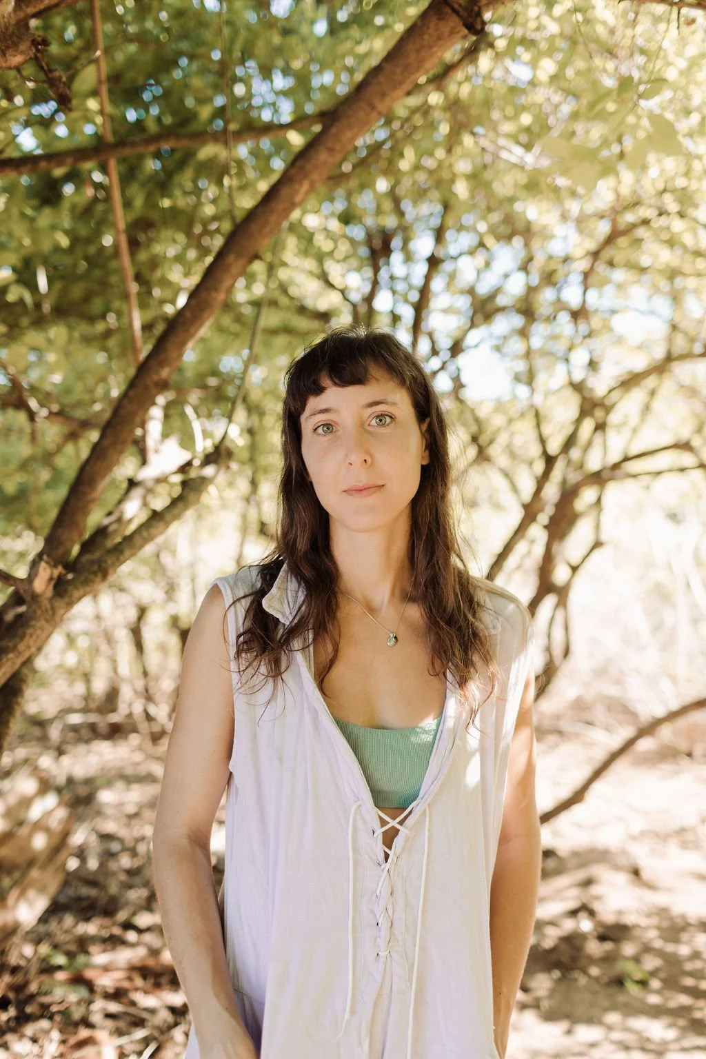 A woman with long, dark hair and light green eyes standing outdoors among trees with green leaves, looking at the camera.