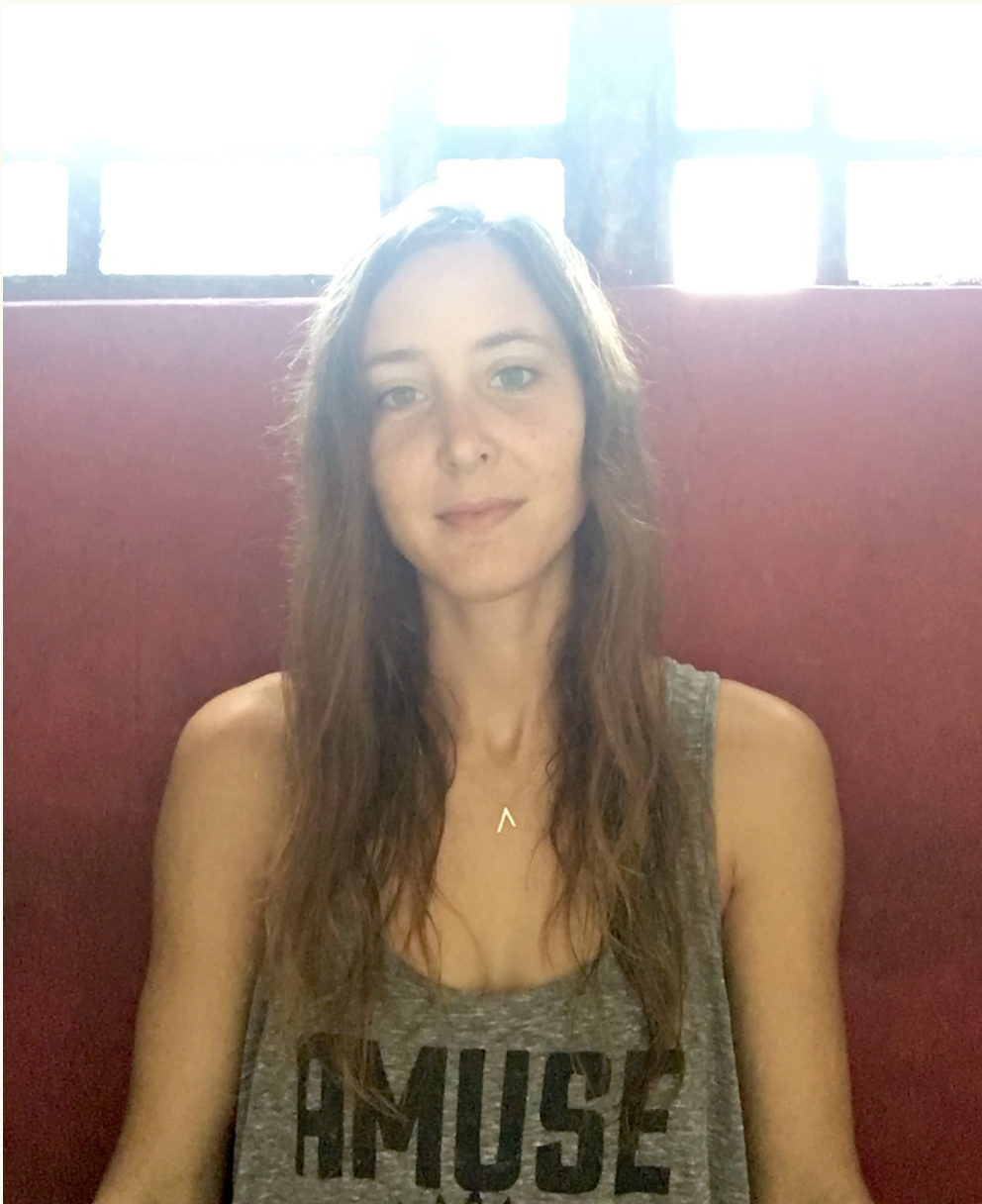 A young woman with light skin and long wavy brown hair, wearing a gray sleeveless top with the word "MUSE" on it, sitting against a red wall with bright windows behind her.