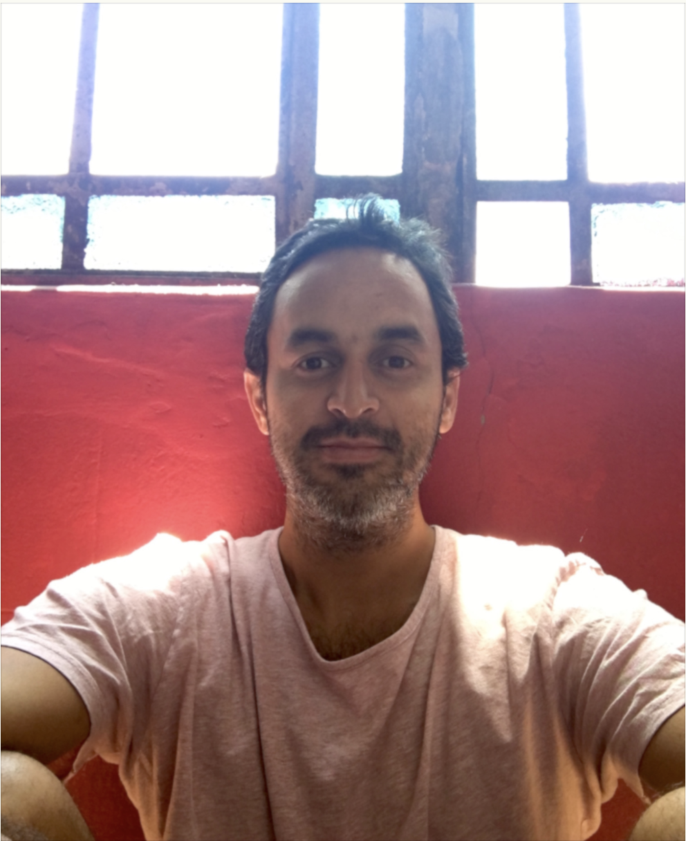 A man with dark hair and a beard, wearing a light-colored t-shirt, sitting in front of a red wall with a window in the background.