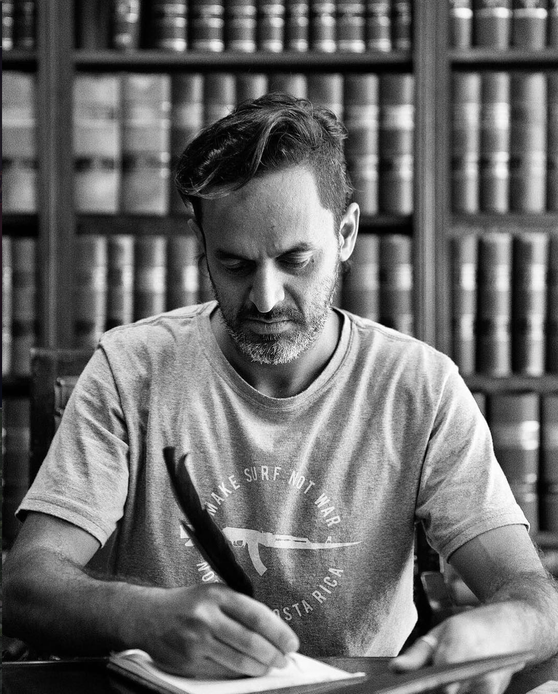 A man with short hair and a beard, wearing a T-shirt, is sitting at a table writing in a notebook, with a backdrop of bookshelves filled with books.