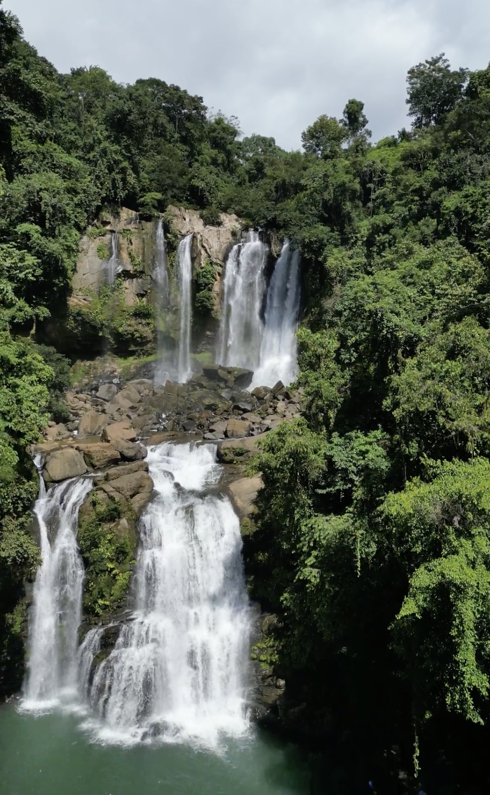 Multiple waterfalls cascading down a lush green mountainside into a pool below, with rocks and dense foliage surrounding the scene.